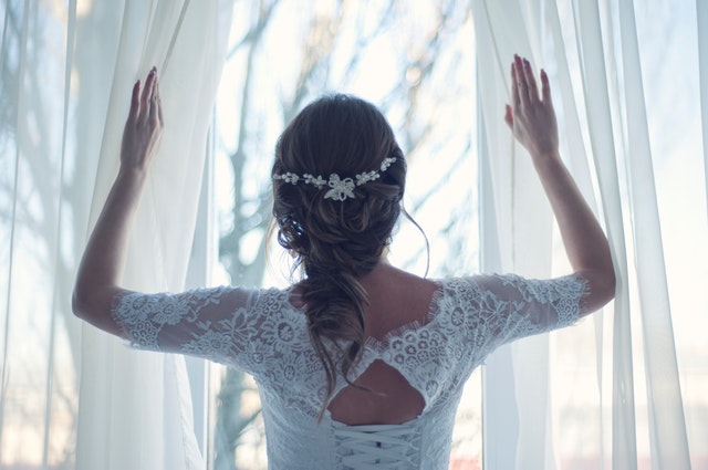 Nevěsta v bílých krajkových šatech stojící u okna